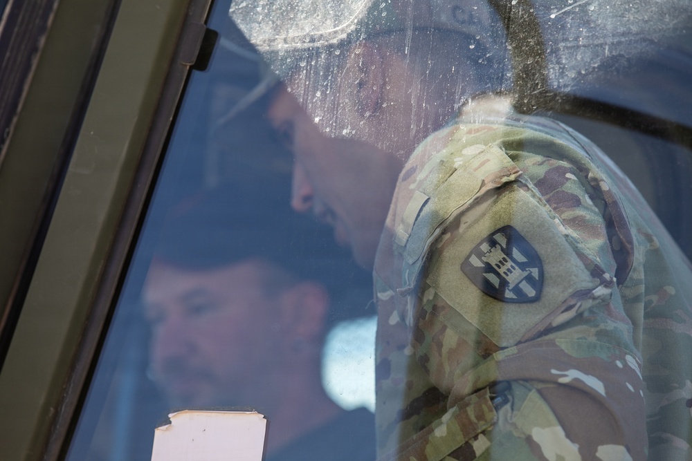 The image shows a military personnel wearing a camouflage uniform (OCP/MultiCam pattern) viewed through a vehicle or building window. A shield-shaped unit patch is visible on the left shoulder of the uniform. The glass surface is dirty and reflective, creating layered visual effects with reflections of additional figures visible.