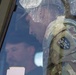 The image shows a military personnel wearing a camouflage uniform (OCP/MultiCam pattern) viewed through a vehicle or building window. A shield-shaped unit patch is visible on the left shoulder of the uniform. The glass surface is dirty and reflective, creating layered visual effects with reflections of additional figures visible.