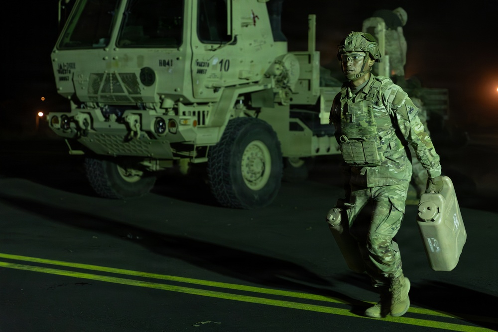 A soldier in full combat gear is photographed at night carrying a military-style jerry can while walking in front of a large tactical military truck. The scene is illuminated by artificial green-tinted lighting, suggesting a nighttime military operation or exercise. The soldier is wearing camouflage uniform, body armor, helmet, and protective eyewear.
