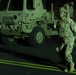 A soldier in full combat gear is photographed at night carrying a military-style jerry can while walking in front of a large tactical military truck. The scene is illuminated by artificial green-tinted lighting, suggesting a nighttime military operation or exercise. The soldier is wearing camouflage uniform, body armor, helmet, and protective eyewear.