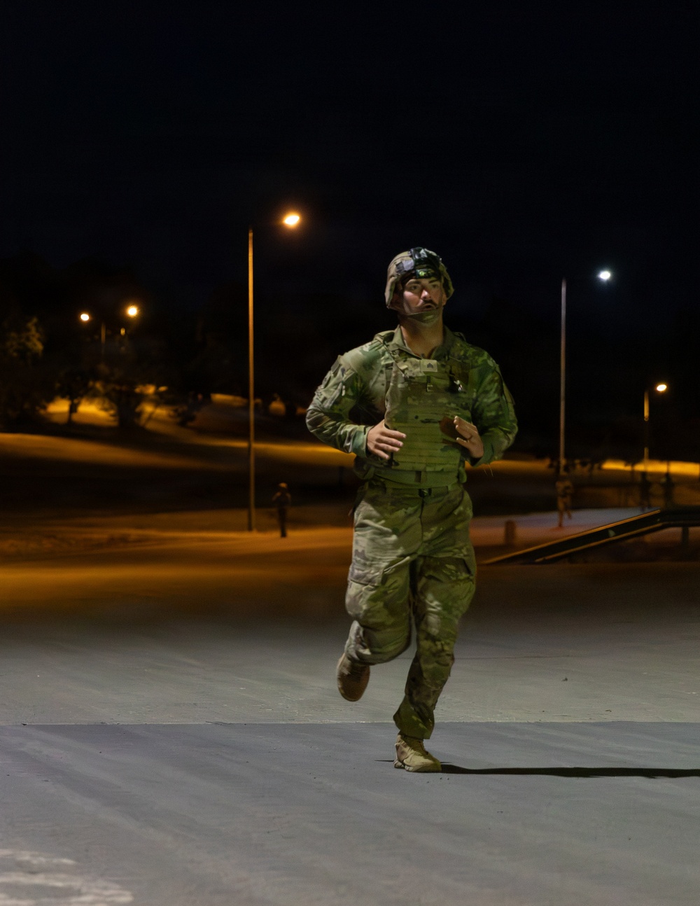 A soldier in full combat gear is running at night on what appears to be a military installation or base. The individual is wearing OCP (Operational Camouflage Pattern) uniform, a tactical vest/plate carrier, and a helmet with night vision mount. The scene is illuminated by street lights casting warm orange and white light across the area.