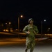 A soldier in full combat gear is running at night on what appears to be a military installation or base. The individual is wearing OCP (Operational Camouflage Pattern) uniform, a tactical vest/plate carrier, and a helmet with night vision mount. The scene is illuminated by street lights casting warm orange and white light across the area.