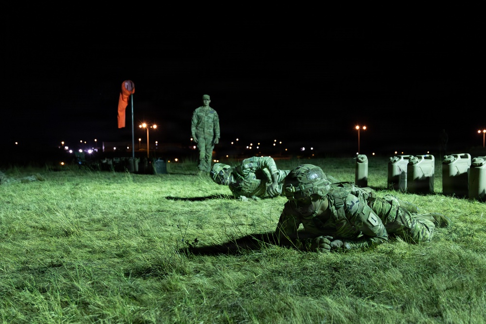 A nighttime military training exercise showing soldiers in full combat gear and camouflage uniforms low-crawling across a grass field. A standing instructor or observer in uniform monitors the exercise from the background. The scene is illuminated by artificial lighting, revealing a structured training lane marked with containers on the right side.
