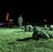 A nighttime military training exercise showing soldiers in full combat gear and camouflage uniforms low-crawling across a grass field. A standing instructor or observer in uniform monitors the exercise from the background. The scene is illuminated by artificial lighting, revealing a structured training lane marked with containers on the right side.