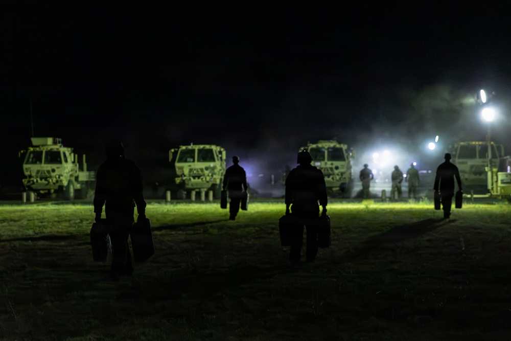 A nighttime military scene showing multiple silhouetted personnel walking toward a line of military tactical vehicles illuminated by artificial lighting. The figures appear to be carrying equipment or cases as they move across an open grassy area. The scene suggests a nighttime operation, exercise, or deployment activity.
