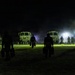 A nighttime military scene showing multiple silhouetted personnel walking toward a line of military tactical vehicles illuminated by artificial lighting. The figures appear to be carrying equipment or cases as they move across an open grassy area. The scene suggests a nighttime operation, exercise, or deployment activity.