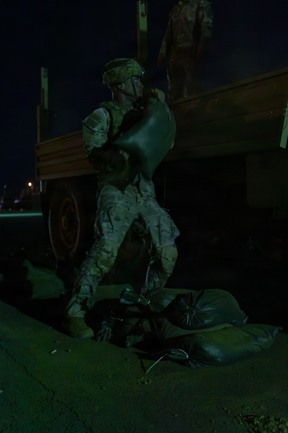 A soldier in full combat uniform and helmet is unloading or stacking sandbags from a military truck at night. The operation appears to be a fortification or flood-control task being conducted under low-light conditions. At least two sandbags are visible on the ground near the soldier's feet.