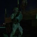 A soldier in full combat uniform and helmet is unloading or stacking sandbags from a military truck at night. The operation appears to be a fortification or flood-control task being conducted under low-light conditions. At least two sandbags are visible on the ground near the soldier's feet.