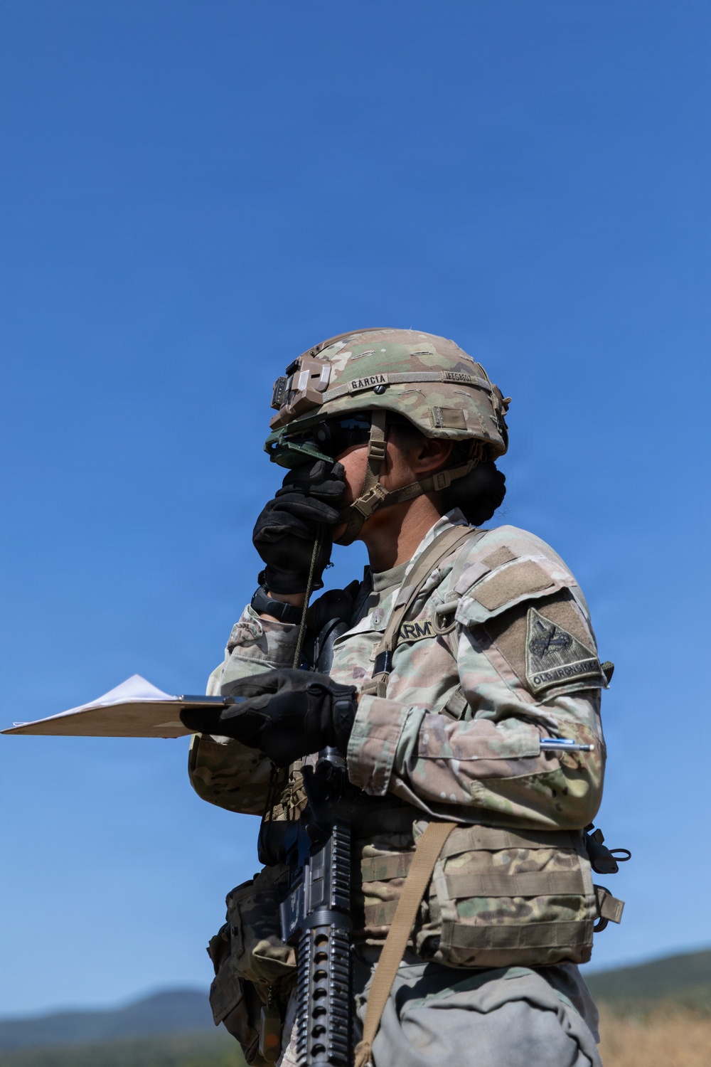 A U.S. Army soldier in full combat gear is standing outdoors under a clear blue sky, holding a radio handset to their face while referencing a clipboard. The soldier is wearing Operational Camouflage Pattern (OCP) uniform with a tactical helmet bearing the name 'GARCIA' and a unit patch reading 'OLD IRONSIDES' (1st Armored Division). The soldier appears to be conducting a radio communication during a field training exercise or operational activity.