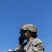 A U.S. Army soldier in full combat gear is standing outdoors under a clear blue sky, holding a radio handset to their face while referencing a clipboard. The soldier is wearing Operational Camouflage Pattern (OCP) uniform with a tactical helmet bearing the name 'GARCIA' and a unit patch reading 'OLD IRONSIDES' (1st Armored Division). The soldier appears to be conducting a radio communication during a field training exercise or operational activity.