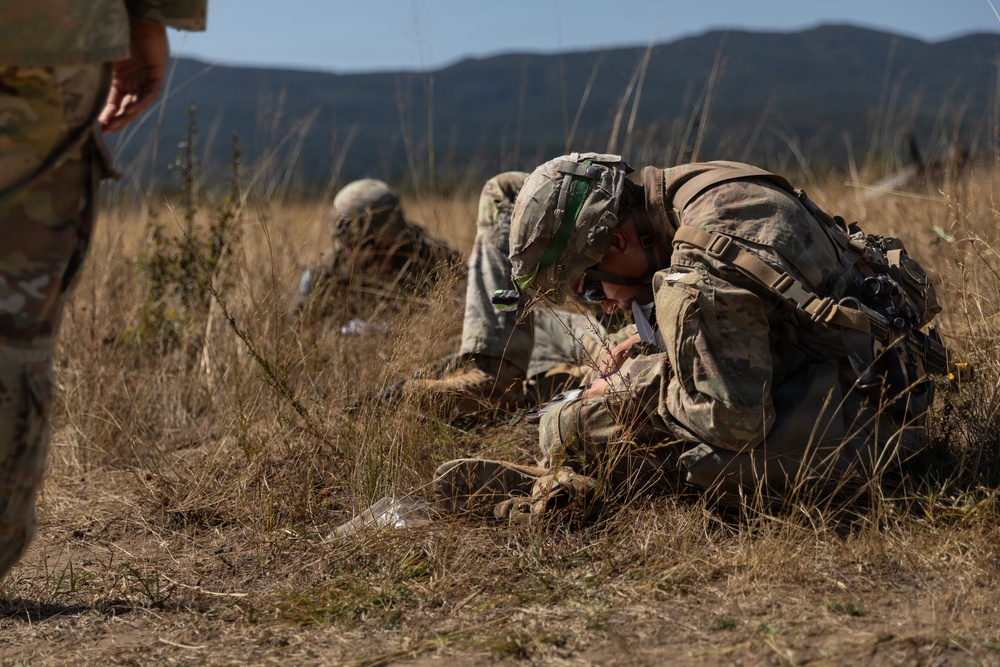 The image shows military personnel in camouflage uniforms and helmets performing what appears to be a combat casualty care or tactical field medicine exercise in an open grassy field. At least three soldiers are visible, with one in the foreground leaning over and administering treatment to a simulated casualty on the ground. The setting appears to be a training environment with mountainous terrain visible in the background.