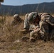 The image shows military personnel in camouflage uniforms and helmets performing what appears to be a combat casualty care or tactical field medicine exercise in an open grassy field. At least three soldiers are visible, with one in the foreground leaning over and administering treatment to a simulated casualty on the ground. The setting appears to be a training environment with mountainous terrain visible in the background.