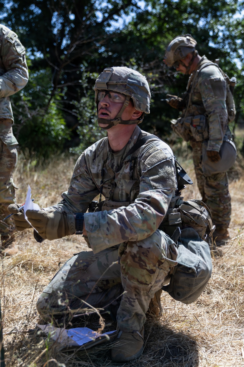 A U.S. Army soldier identified by the name tape 'ATKINSON' on his helmet is kneeling in a field, holding papers and looking upward, apparently during a field training exercise. He is wearing MultiCam (OCP) camouflage uniform, tactical gear, protective eyewear, and an Advanced Combat Helmet. At least two other soldiers in similar gear are visible in the background amid dry grass and trees.