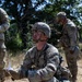 A U.S. Army soldier identified by the name tape 'ATKINSON' on his helmet is kneeling in a field, holding papers and looking upward, apparently during a field training exercise. He is wearing MultiCam (OCP) camouflage uniform, tactical gear, protective eyewear, and an Advanced Combat Helmet. At least two other soldiers in similar gear are visible in the background amid dry grass and trees.
