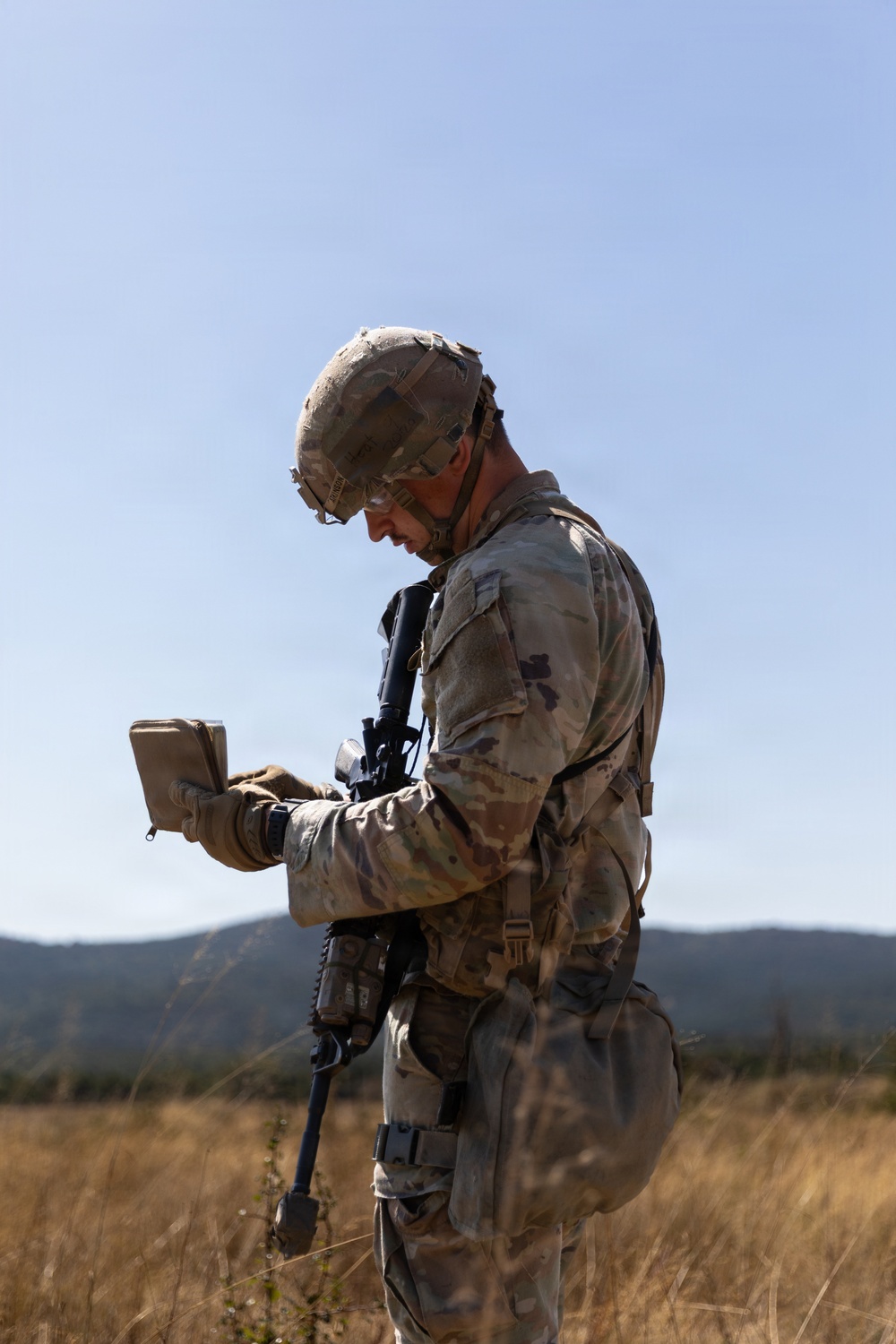 A soldier in full combat gear stands in an open dry grassland field, consulting what appears to be a small notebook or field manual. The individual is equipped with a rifle, tactical helmet, body armor, and multicam/OCP camouflage uniform. Rolling hills and a clear blue sky form the background, suggesting a training exercise or field operation.