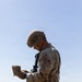 A soldier in full combat gear stands in an open dry grassland field, consulting what appears to be a small notebook or field manual. The individual is equipped with a rifle, tactical helmet, body armor, and multicam/OCP camouflage uniform. Rolling hills and a clear blue sky form the background, suggesting a training exercise or field operation.