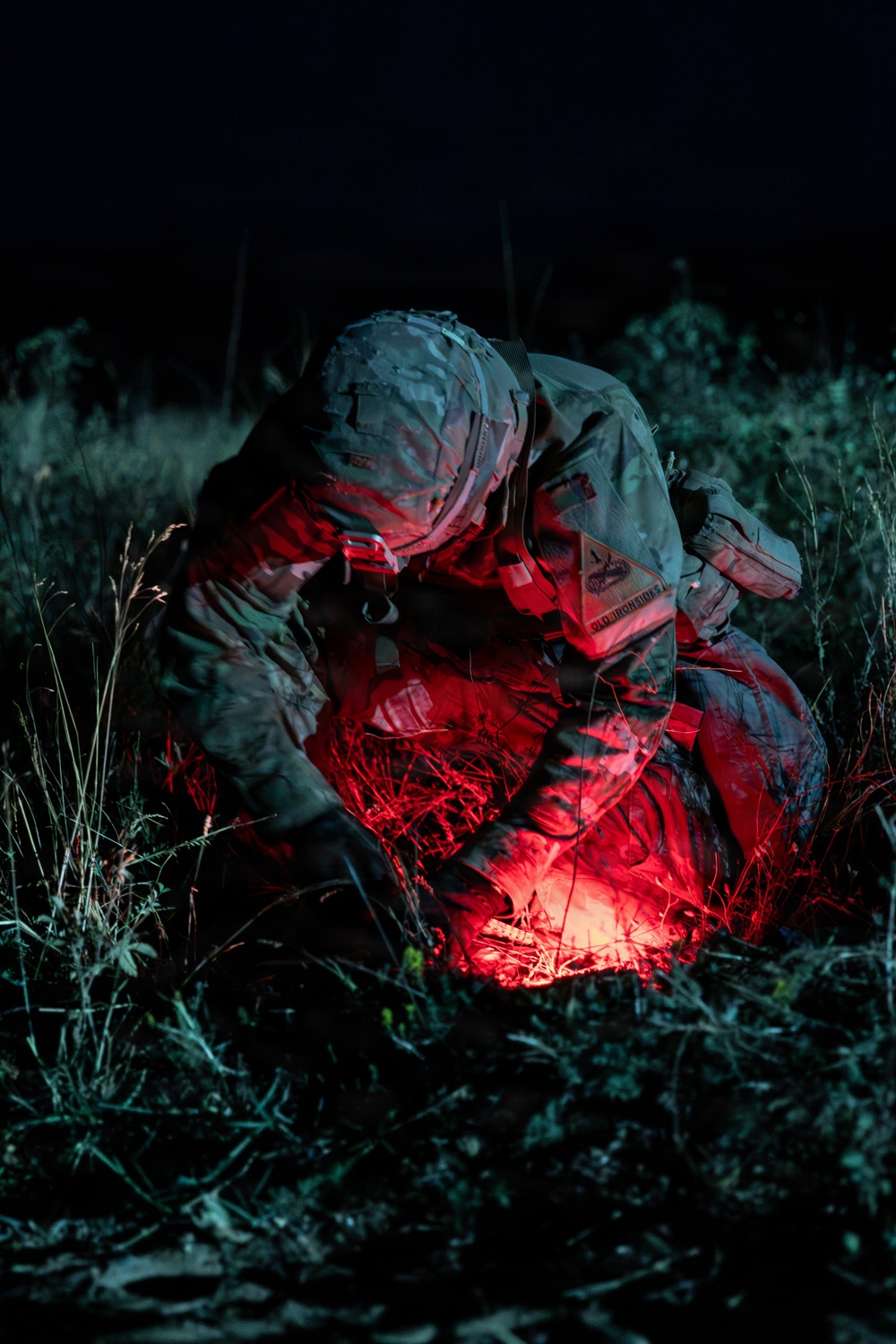 A military personnel in full combat gear is crouched over a casualty or object on the ground at night, illuminated by a red tactical light. The individual appears to be performing a medical treatment or field procedure, suggested by visible medical pouches and equipment. The scene is set in an outdoor grassy area under complete darkness.