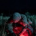 A military personnel in full combat gear is crouched over a casualty or object on the ground at night, illuminated by a red tactical light. The individual appears to be performing a medical treatment or field procedure, suggested by visible medical pouches and equipment. The scene is set in an outdoor grassy area under complete darkness.