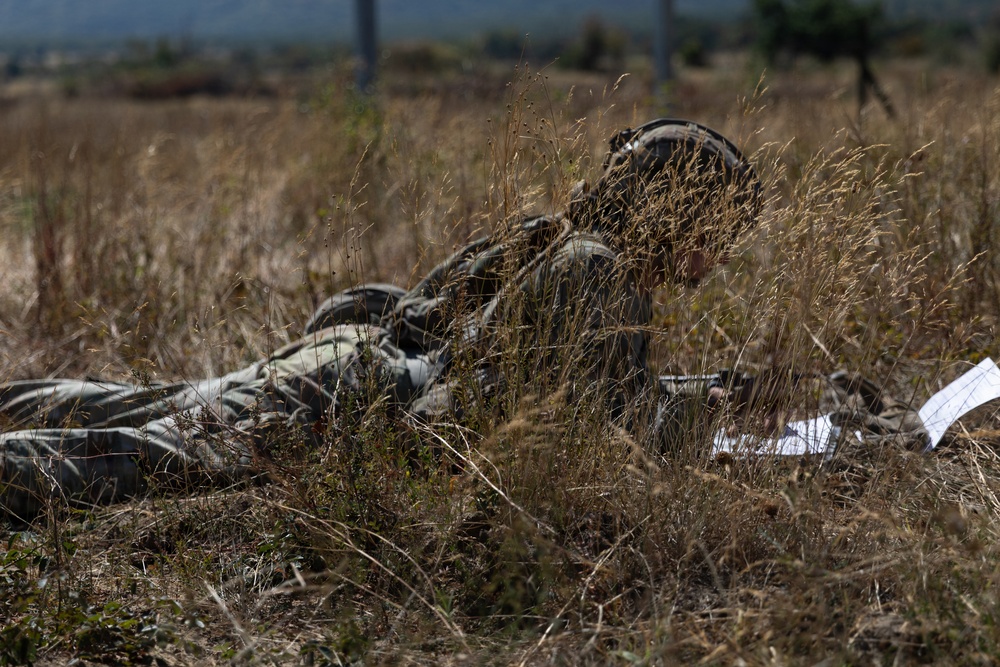 A soldier or sniper in camouflage gear is lying prone in a dry grassy field, blending into the surrounding vegetation. The individual appears to be in a concealed observation or firing position, consistent with military training or tactical operations. A white document or paper is visible to the right of the figure.