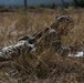 A soldier or sniper in camouflage gear is lying prone in a dry grassy field, blending into the surrounding vegetation. The individual appears to be in a concealed observation or firing position, consistent with military training or tactical operations. A white document or paper is visible to the right of the figure.