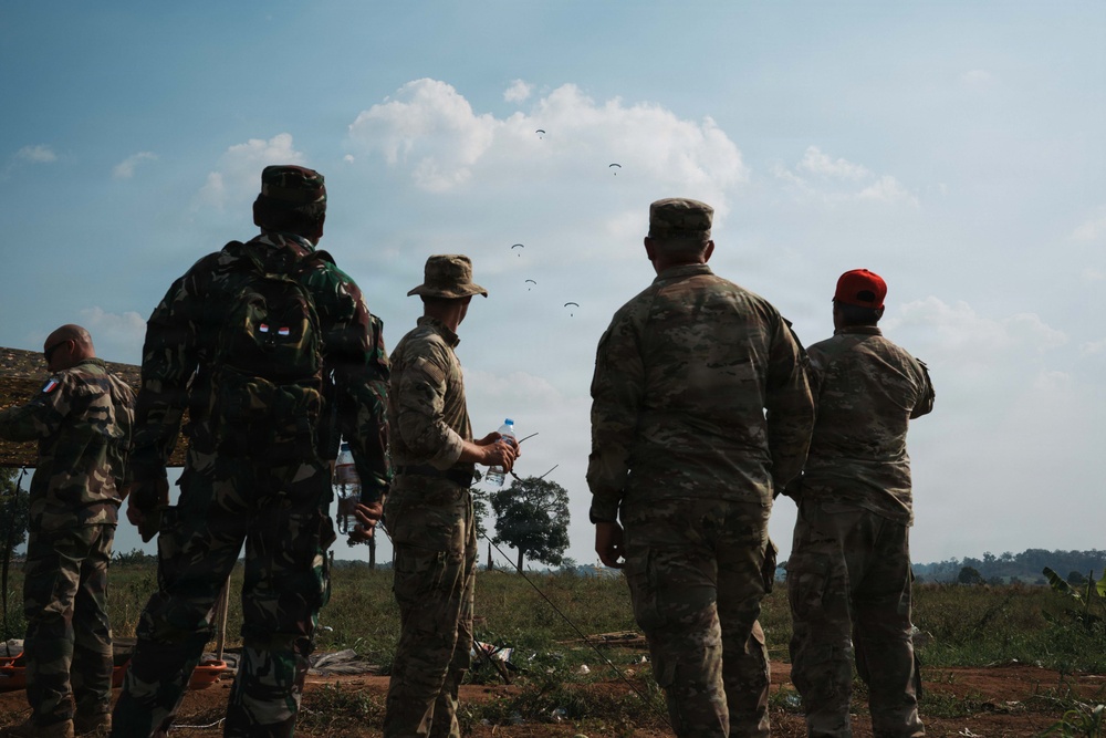 A group of military personnel from multiple nations are observed standing in an open field, watching what appear to be paratroopers or parachutists descending in the sky. The soldiers are wearing different camouflage uniforms, with at least one Indonesian flag patch visible on a uniform. The scene suggests a multinational military exercise or joint training operation.