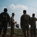 A group of military personnel from multiple nations are observed standing in an open field, watching what appear to be paratroopers or parachutists descending in the sky. The soldiers are wearing different camouflage uniforms, with at least one Indonesian flag patch visible on a uniform. The scene suggests a multinational military exercise or joint training operation.