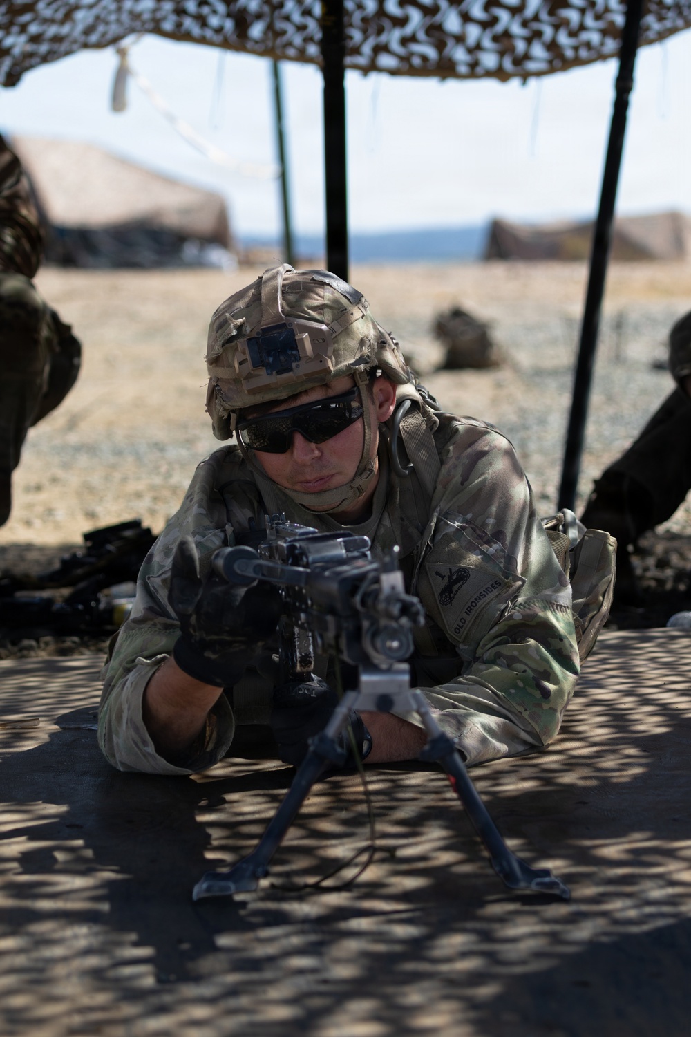 A U.S. Army soldier in full combat gear is lying in a prone position behind a bipod-mounted weapon system under a camouflage netting canopy. The soldier is wearing Operational Camouflage Pattern (OCP) uniform with a patch reading 'OLD IRONSIDES' (1st Armored Division) visible on the right sleeve. The setting appears to be a desert training environment.