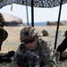 A U.S. Army soldier in full combat gear is lying in a prone position behind a bipod-mounted weapon system under a camouflage netting canopy. The soldier is wearing Operational Camouflage Pattern (OCP) uniform with a patch reading 'OLD IRONSIDES' (1st Armored Division) visible on the right sleeve. The setting appears to be a desert training environment.
