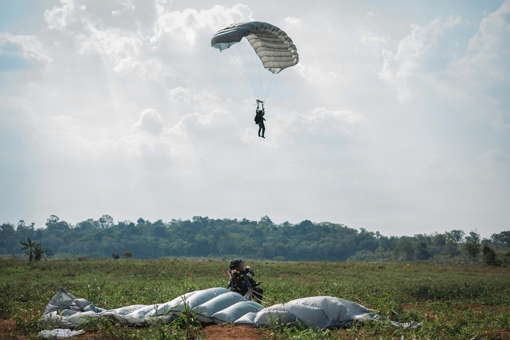 A military parachute operation is underway over an open field with a treeline in the background. One paratrooper is still descending under a gray/silver ram-air parachute canopy, while another has already landed and is crouching near a collapsed parachute on the ground. The setting appears to be a rural drop zone, likely in a tropical or subtropical region based on the vegetation.