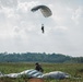 A military parachute operation is underway over an open field with a treeline in the background. One paratrooper is still descending under a gray/silver ram-air parachute canopy, while another has already landed and is crouching near a collapsed parachute on the ground. The setting appears to be a rural drop zone, likely in a tropical or subtropical region based on the vegetation.