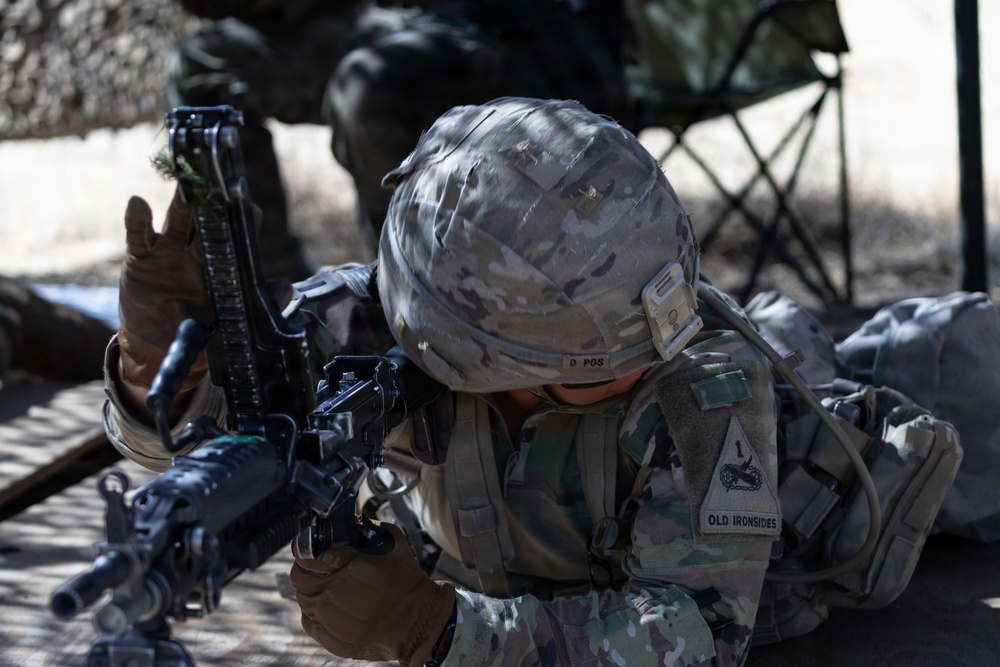 A U.S. Army soldier in full combat gear is in a prone or crouched position handling an assault rifle, appearing to perform a weapons check or malfunction drill. The soldier's uniform patch reads 'OLD IRONSIDES,' identifying them as a member of the 1st Armored Division. The setting appears to be a training range or field exercise environment.