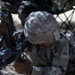 A U.S. Army soldier in full combat gear is in a prone or crouched position handling an assault rifle, appearing to perform a weapons check or malfunction drill. The soldier's uniform patch reads 'OLD IRONSIDES,' identifying them as a member of the 1st Armored Division. The setting appears to be a training range or field exercise environment.