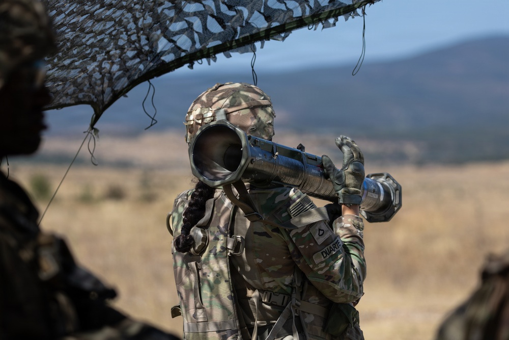A U.S. Army soldier identified by the name tape 'DUARTE' is shouldering and aiming a shoulder-fired rocket launcher, likely an AT4 or similar anti-armor weapon, during what appears to be a field training exercise. The soldier is wearing MultiCam (OCP) camouflage uniform and helmet, with a U.S. flag patch visible on the right sleeve. The setting is an open, arid training range with mountainous terrain in the background.
