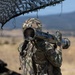 A U.S. Army soldier identified by the name tape 'DUARTE' is shouldering and aiming a shoulder-fired rocket launcher, likely an AT4 or similar anti-armor weapon, during what appears to be a field training exercise. The soldier is wearing MultiCam (OCP) camouflage uniform and helmet, with a U.S. flag patch visible on the right sleeve. The setting is an open, arid training range with mountainous terrain in the background.