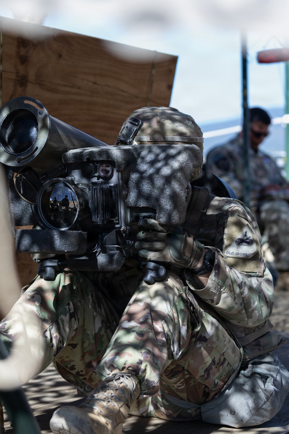 A U.S. Army soldier in MultiCam/OCP camouflage uniform is aiming through the sight of what appears to be a Javelin or similar man-portable anti-tank guided missile (ATGM) system. The soldier's shoulder patch reads 'OLD IRONSIDES,' identifying them as a member of the 1st Armored Division. A second soldier in uniform is visible in the background, and the setting appears to be a training or demonstration environment.