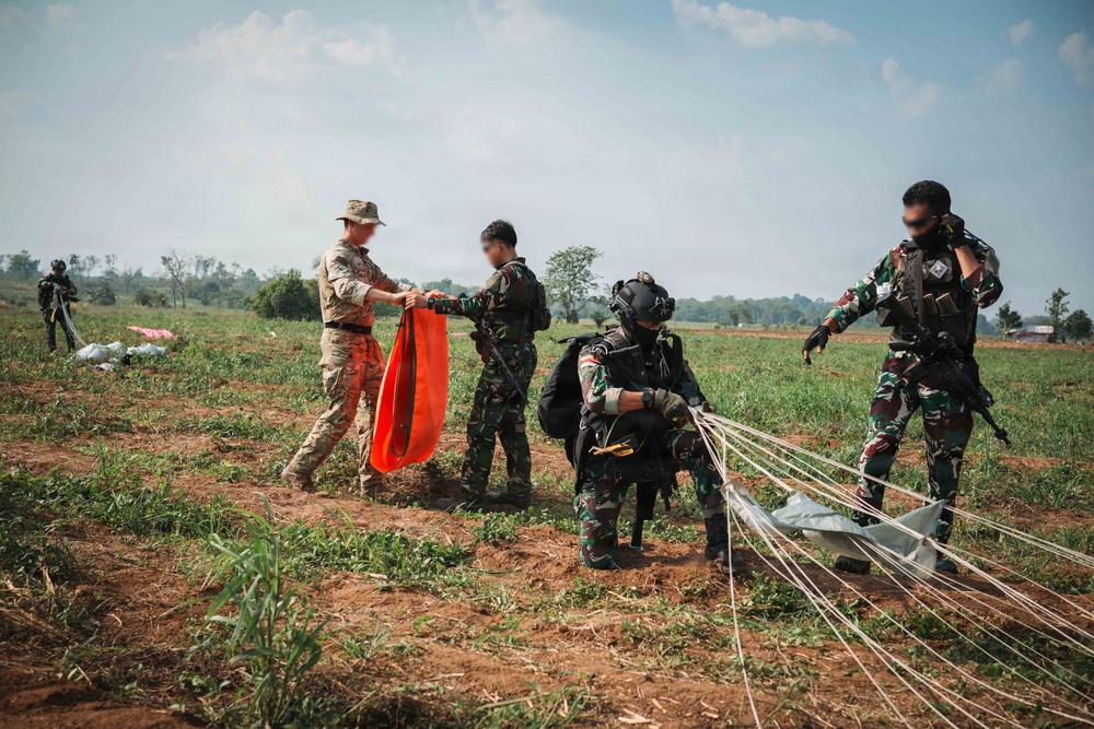 Military personnel are gathered on an open field following what appears to be a parachute landing exercise. One soldier in tan/multicam uniform is holding an orange signal panel, while others in camouflage uniforms are managing parachute lines and equipment. The scene suggests a joint military training operation on a drop zone.