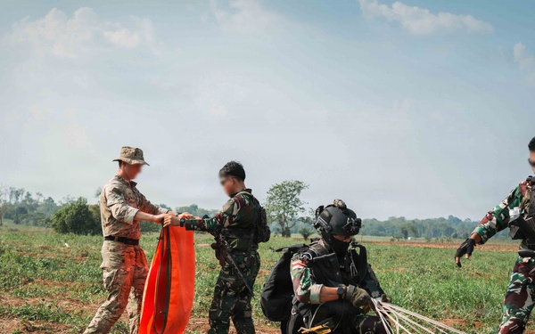 Military personnel are gathered on an open field following what appears to be a parachute landing exercise. One soldier in tan/multicam uniform is holding an orange signal panel, while others in camouflage uniforms are managing parachute lines and equipment. The scene suggests a joint military training operation on a drop zone.