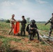 Military personnel are gathered on an open field following what appears to be a parachute landing exercise. One soldier in tan/multicam uniform is holding an orange signal panel, while others in camouflage uniforms are managing parachute lines and equipment. The scene suggests a joint military training operation on a drop zone.
