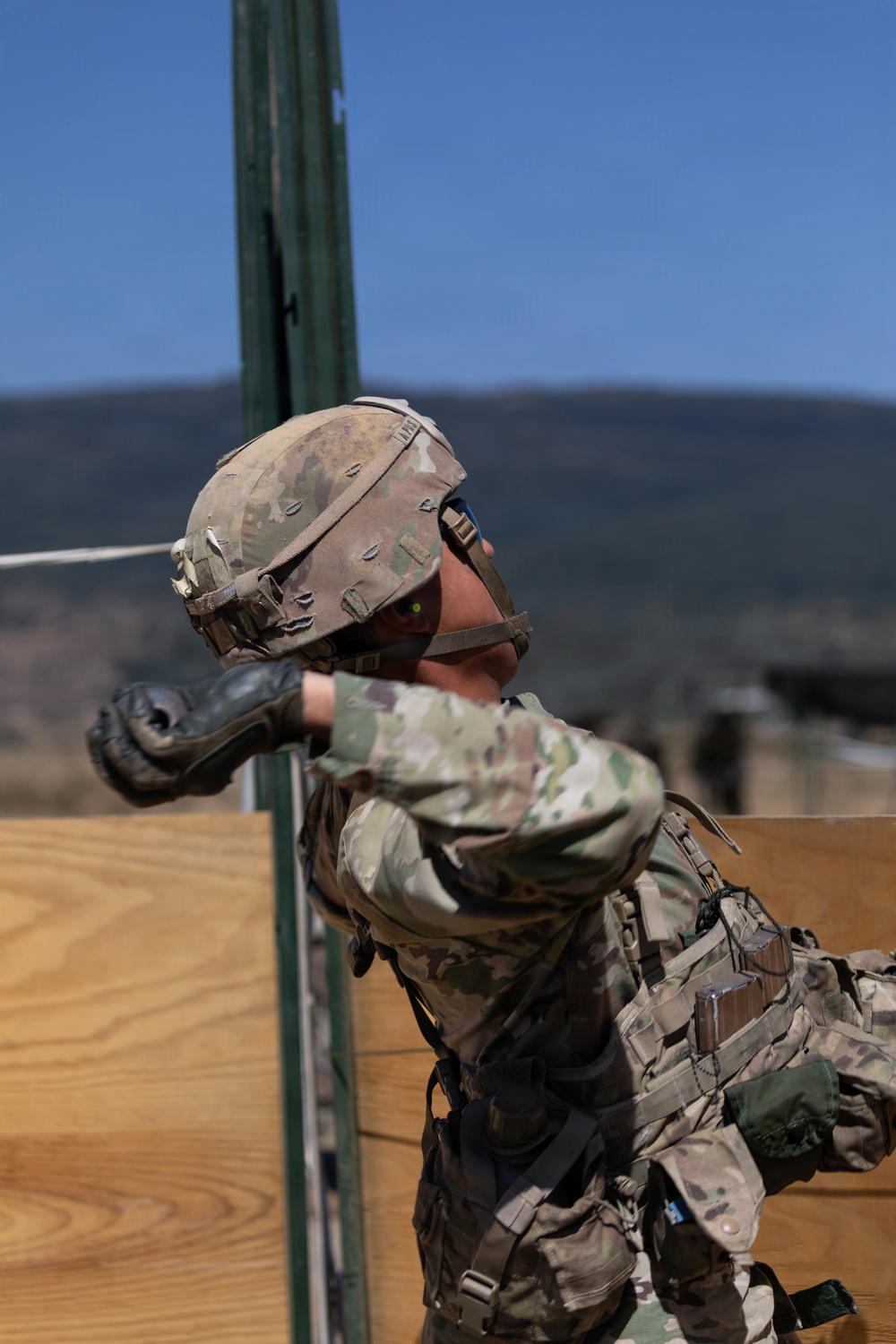 A soldier in full combat gear and MultiCam/OCP camouflage uniform is engaged in what appears to be a military training exercise at an outdoor range or obstacle course. The soldier is wearing a tactical helmet with a cover, black gloves, and a plate carrier with pouches, and appears to be navigating or pushing through a wooden barrier structure. The setting is a clear-sky outdoor environment with mountainous terrain visible in the background.