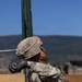 A soldier in full combat gear and MultiCam/OCP camouflage uniform is engaged in what appears to be a military training exercise at an outdoor range or obstacle course. The soldier is wearing a tactical helmet with a cover, black gloves, and a plate carrier with pouches, and appears to be navigating or pushing through a wooden barrier structure. The setting is a clear-sky outdoor environment with mountainous terrain visible in the background.