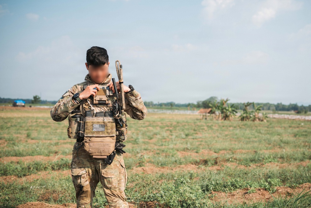 A soldier in full tactical gear stands in an open field with sparse vegetation. The individual is wearing MultiCam camouflage uniform with a plate carrier vest and carrying a rifle over one shoulder. The face has been deliberately obscured/blurred, suggesting identity protection.