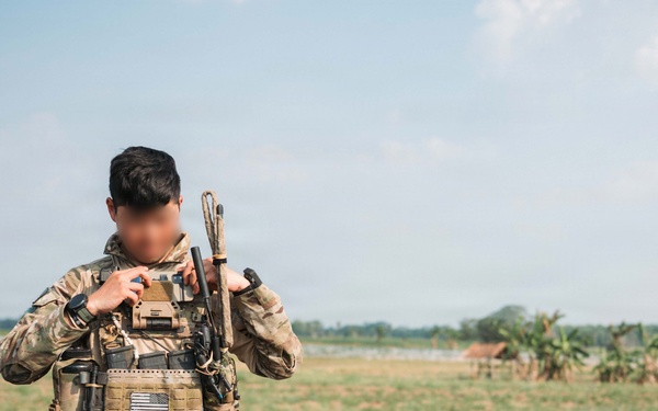 A soldier in full tactical gear stands in an open field with sparse vegetation. The individual is wearing MultiCam camouflage uniform with a plate carrier vest and carrying a rifle over one shoulder. The face has been deliberately obscured/blurred, suggesting identity protection.