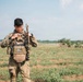 A soldier in full tactical gear stands in an open field with sparse vegetation. The individual is wearing MultiCam camouflage uniform with a plate carrier vest and carrying a rifle over one shoulder. The face has been deliberately obscured/blurred, suggesting identity protection.