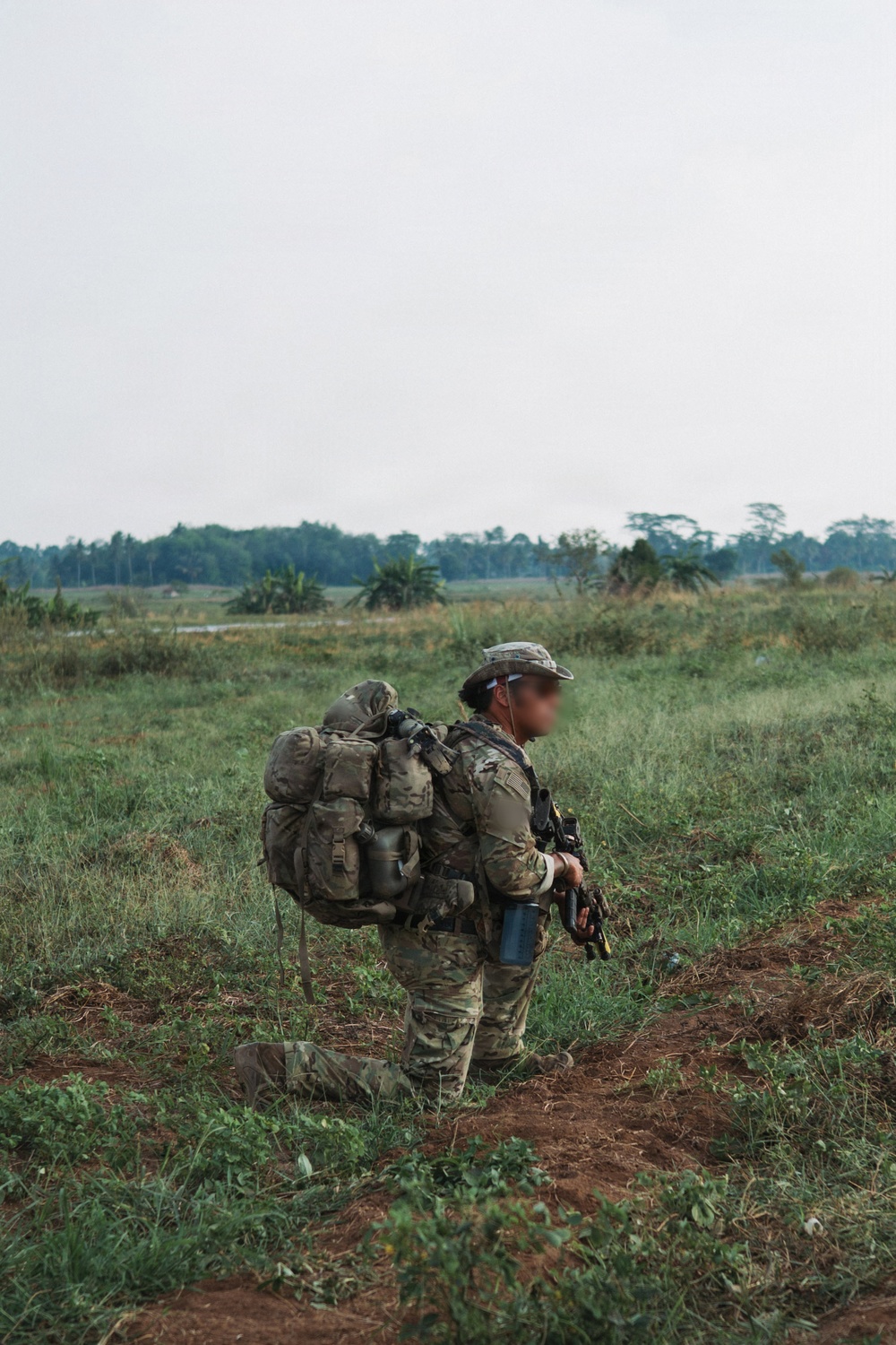 A soldier in full combat gear is kneeling or walking on a dirt path in an open grassy field. The individual is wearing multicam camouflage uniform and carrying a heavily loaded rucksack along with additional equipment. The setting appears to be a tropical or subtropical environment based on the vegetation and tree line in the background.