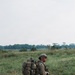 A soldier in full combat gear is kneeling or walking on a dirt path in an open grassy field. The individual is wearing multicam camouflage uniform and carrying a heavily loaded rucksack along with additional equipment. The setting appears to be a tropical or subtropical environment based on the vegetation and tree line in the background.