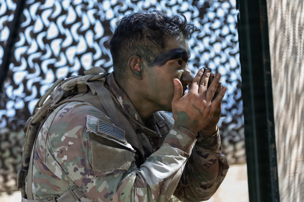 A U.S. Army soldier in full OCP (Operational Camouflage Pattern) uniform is applying camouflage face paint using their hands. The soldier wears a tactical vest and has a U.S. flag patch visible on the left sleeve. The background features a camouflage netting screen, suggesting a field or training environment.