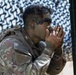 A U.S. Army soldier in full OCP (Operational Camouflage Pattern) uniform is applying camouflage face paint using their hands. The soldier wears a tactical vest and has a U.S. flag patch visible on the left sleeve. The background features a camouflage netting screen, suggesting a field or training environment.