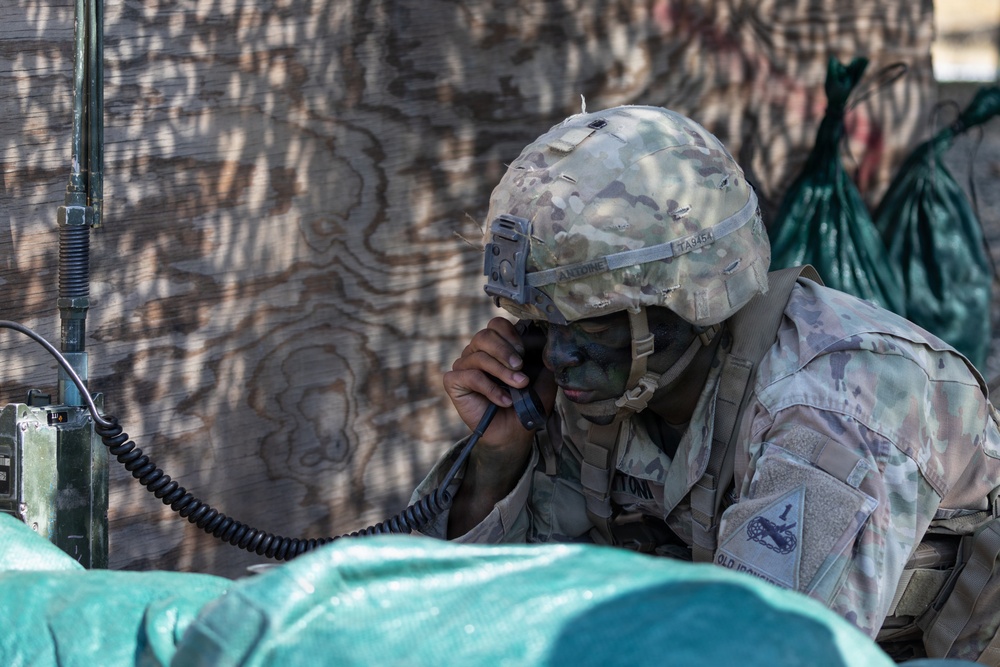A U.S. Army soldier wearing Operational Camouflage Pattern (OCP) uniform and helmet is using a military radio handset, likely during a field training exercise. The soldier has face camouflage paint applied and wears a patch identifying the 1st Cavalry Division ('Old Ironsides'). The soldier appears to be in a defensive or communications position near a plywood structure.