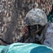 A U.S. Army soldier wearing Operational Camouflage Pattern (OCP) uniform and helmet is using a military radio handset, likely during a field training exercise. The soldier has face camouflage paint applied and wears a patch identifying the 1st Cavalry Division ('Old Ironsides'). The soldier appears to be in a defensive or communications position near a plywood structure.