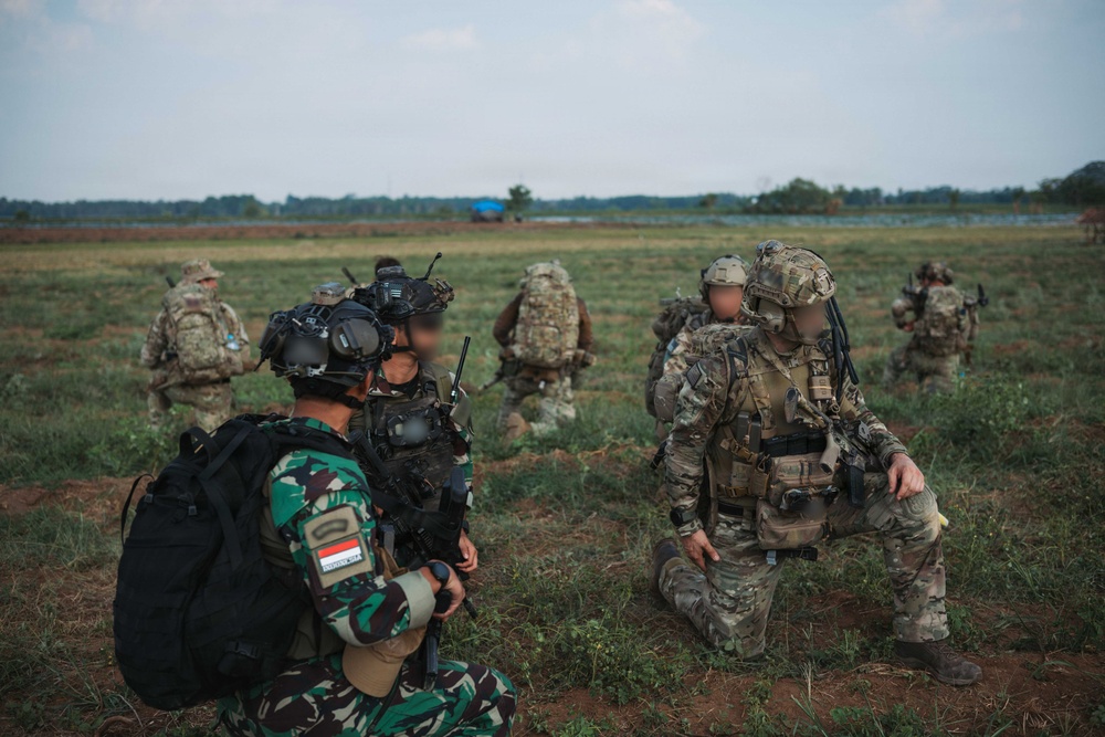 The image shows a joint military exercise or operation involving soldiers from at least two nations in an open field environment. A soldier in the foreground wears an Indonesian flag patch (red and white) on green camouflage uniform, while others wear MultiCam/OCP pattern uniforms consistent with Western military forces. Faces have been digitally blurred, suggesting operational security considerations.